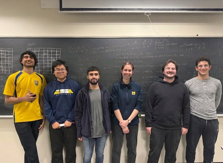 Six people stand in front of a large chalkboard filled with handwritten math equations and grids.Some wear University of Michigan and math-themed apparel.The classroom setting emphasizes teamwork and academic collaboration.