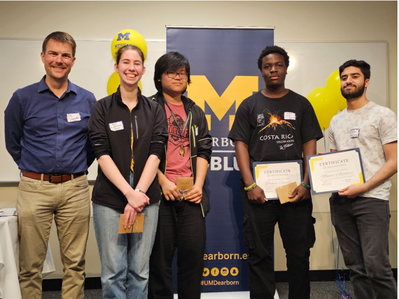 Five people stand in front of a UM-Dearborn banner and yellow balloons.Two individuals hold certificates, and others hold envelope awards.The setting suggests a recognition or award ceremony, with university branding visible.