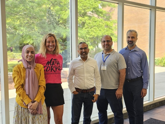 Five people stand in a row indoors near large windows.  One person wears a bright pink "Math Corps" shirt, and another has a yellow jacket with a light scarf.  Trees and brick buildings are visible outside the window in the background.