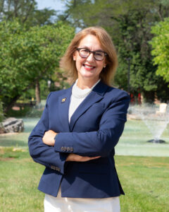 Person standing outdoors with arms crossed, wearing a navy blazer and white shirt.Background includes green trees and a fountain. Bright, sunny day.