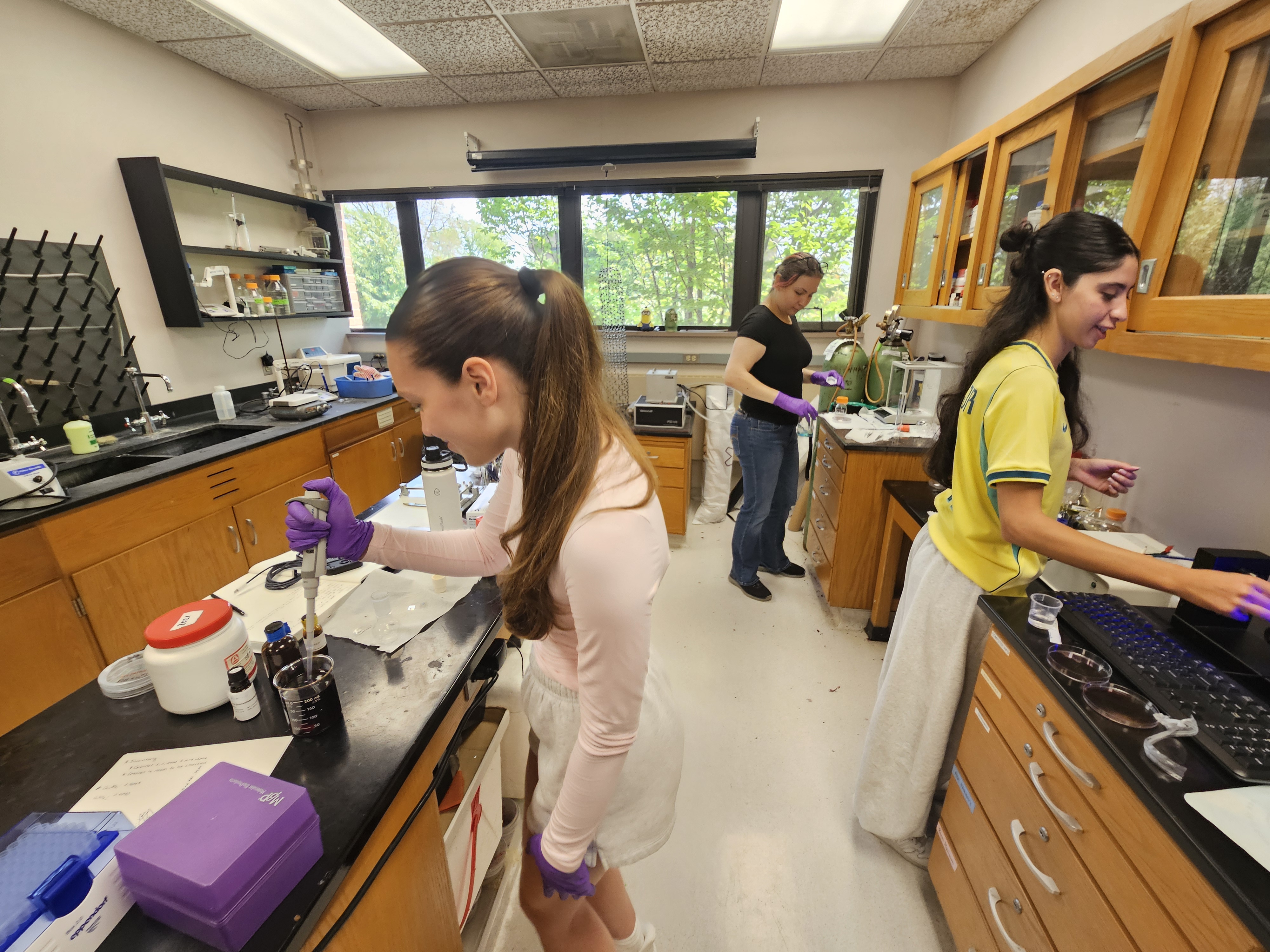 three women work in a lab setting, the left in a pink shirt holding a pipette instrument, the middle background wearing a black shirt and jeans working with chemicals at a station, and the third on the right in a yellow shirt working at a computer microscope station