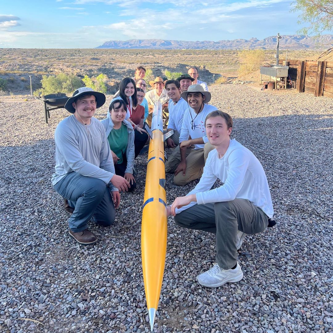 Members of the UM-Dearborn rocket team pose for a photo with their rocket in the New Mexico desert, with mountains in the background.