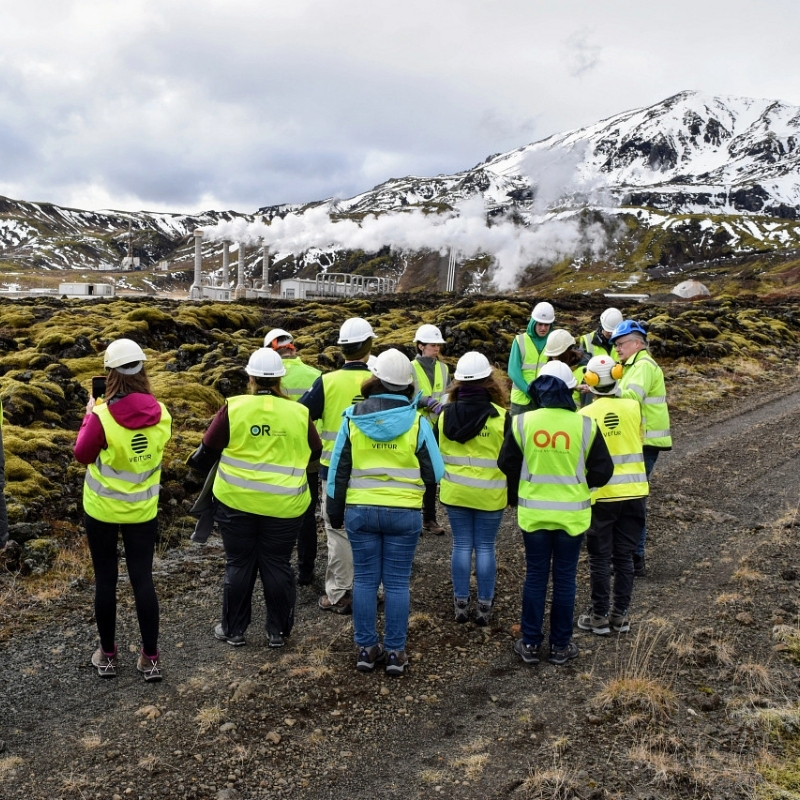 Group of people in safety vests and hard hats standing outdoors near a geothermal power plant in a mountainous landscape.
