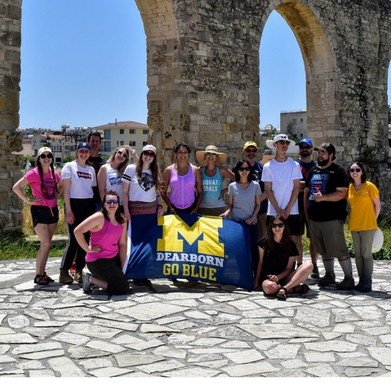 Large group posing with a University of Michigan–Dearborn “Go Blue” banner in front of historic stone arches.