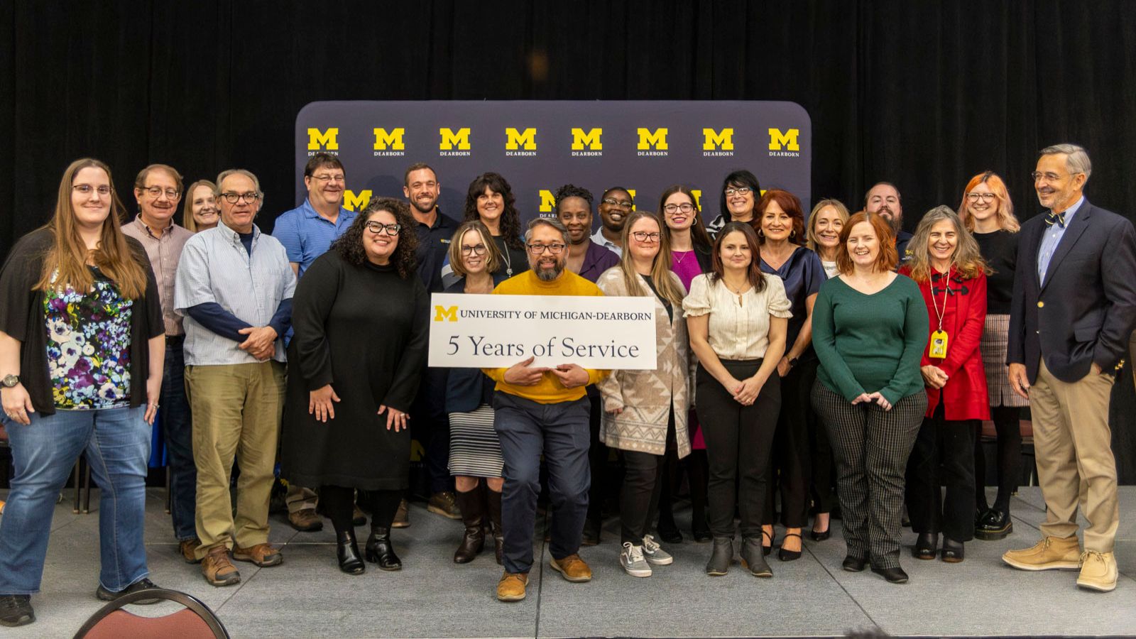 Staff receiving recognition for 5-years of service pictured on stage with Chancellor Grasso in front of a UM-Dearborn background