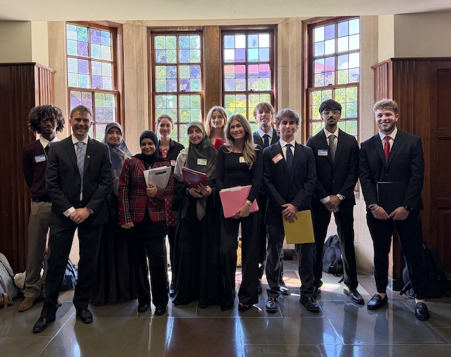 A large, diverse group of students poses for a formal photo.Many wear suits, name tags, and hold folders or papers.They stand in a wood-paneled hall with tall, colorful windows.