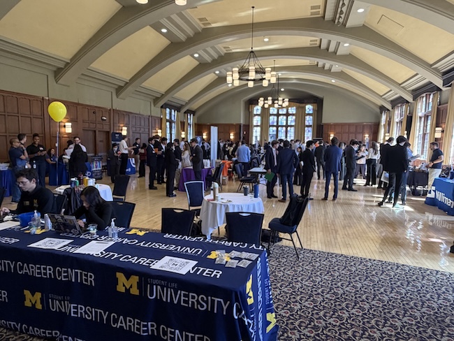 A crowded university career fair fills a large, high-ceilinged hall.Students gather around tables staffed by employers and organizations.University Career Center tablecloths and a yellow balloon appear in front.
