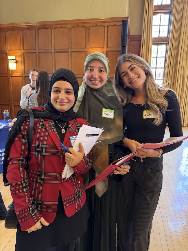 Three smiling students pose closely together in a wood-paneled hall.Three wear hijabs, and two wear coordinated red plaid blazers.They hold papers and folders and wear handwritten name tags.
