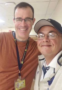 Selfie of two smiling men indoors. The man on the left wears a brown t-shirt and a dark blue lanyard reading "TRANSPLANT CENTER" with a University of Michigan ID badge for Seth A. Waits, MD. The man on the right wears glasses, a baseball cap, and a white Under Armour jacket.