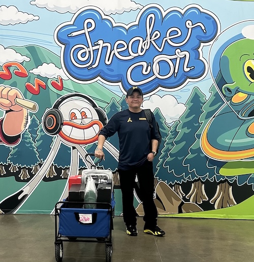 A man in a dark long-sleeve shirt, dark pants, and a Michigan hat stands next to a utility wagon filled with shoe boxes in front of a colorful "Sneaker Con" backdrop.   