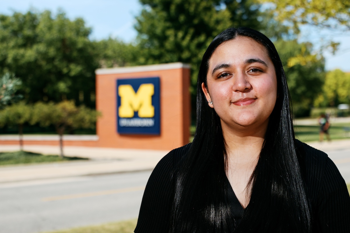 A young woman with long dark hair smiling in front of a University of Michigan-Dearborn sign.
