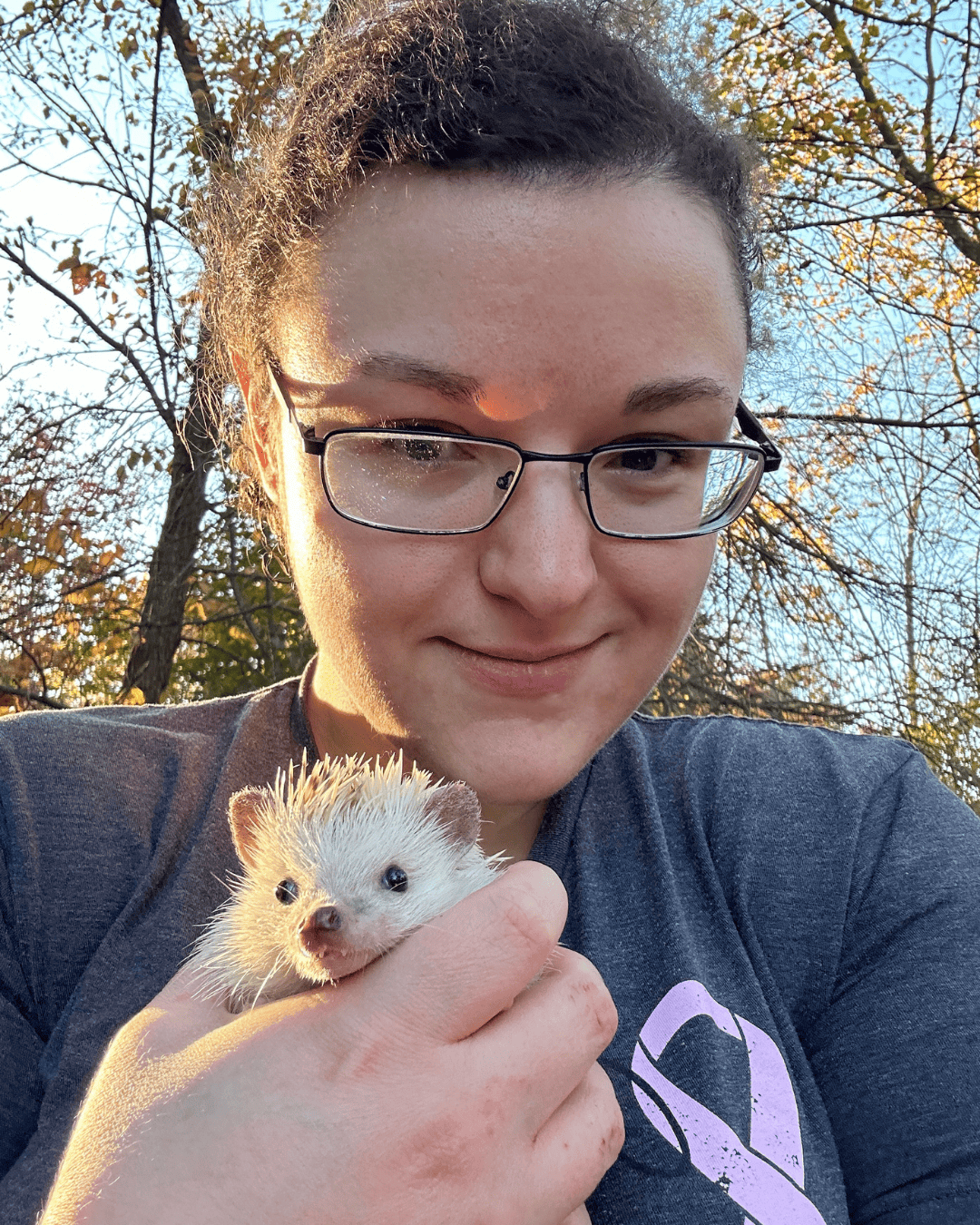 shot of women with brunette hair and black-rimmed glasses outside in front of yellow-leaved trees holding a small hedgehog in her hand