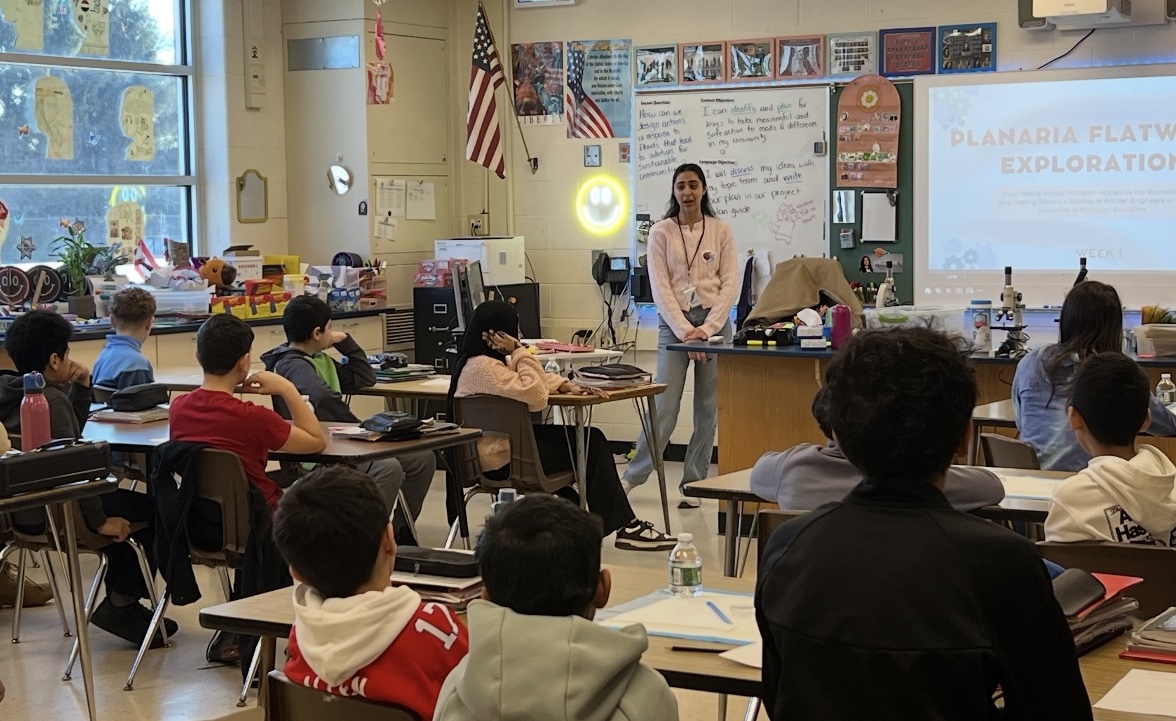 A presenter stands in front of a classroom of seated students giving a presentation titled "Planaria Flatworm Exploration" projected on a screen behind her.