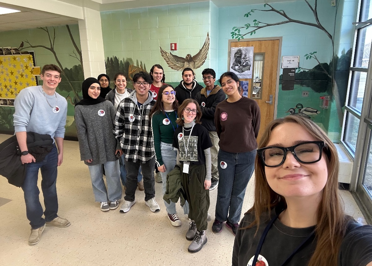 A group selfie of students posing together in a school hallway painted with nature murals, wearing matching pin buttons on their tops.   