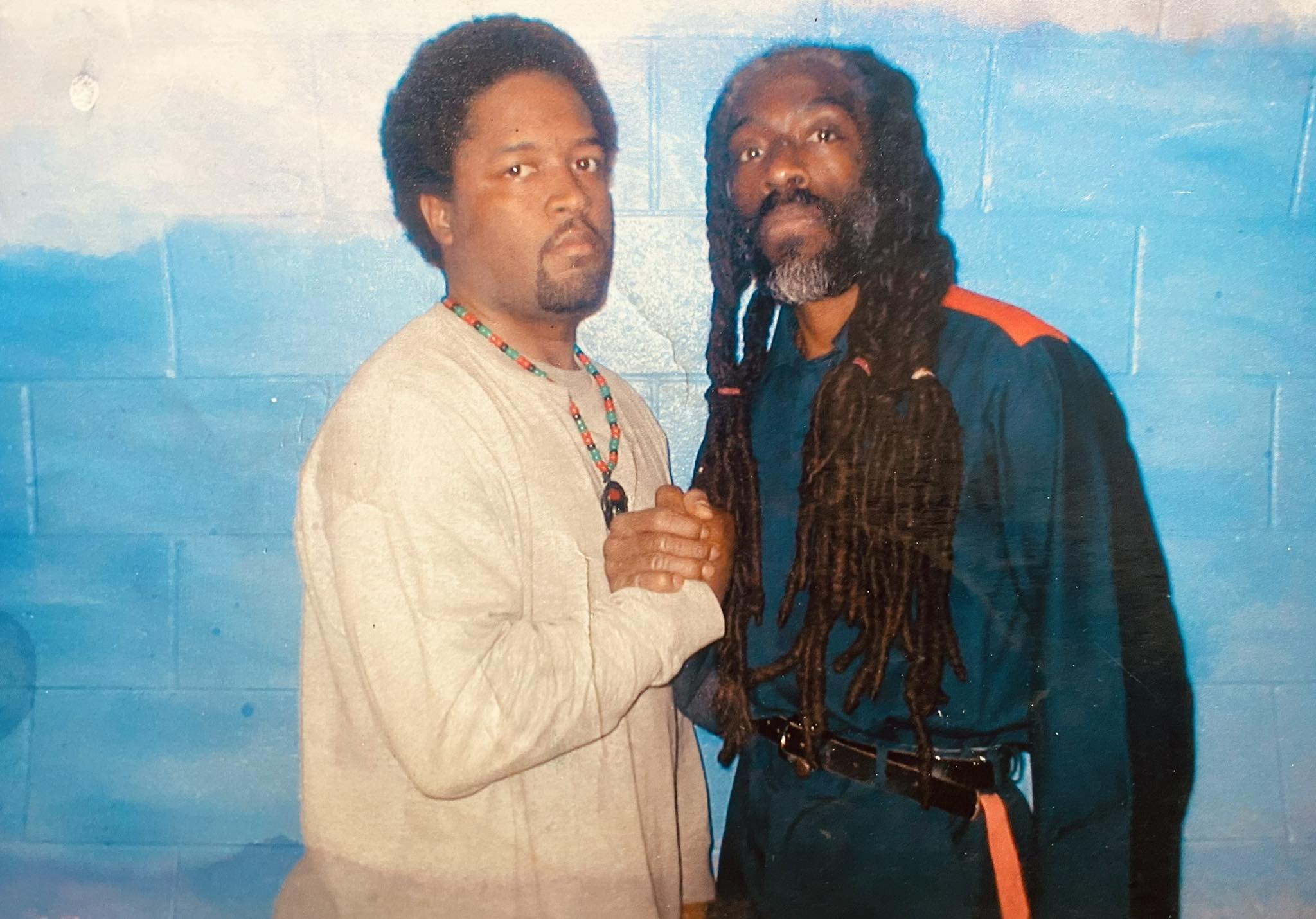 A man with long dreadlocks and his adult son clasp hands while standing for a portrait 
