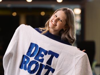 A student entrepreneur holds up a white sweatshirt with the word "DETROIT" in large blue letters while smiling at the camera.