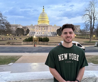 a man stands in front of the capitol building with a blue cloudy sky wearing a "new york" shirt