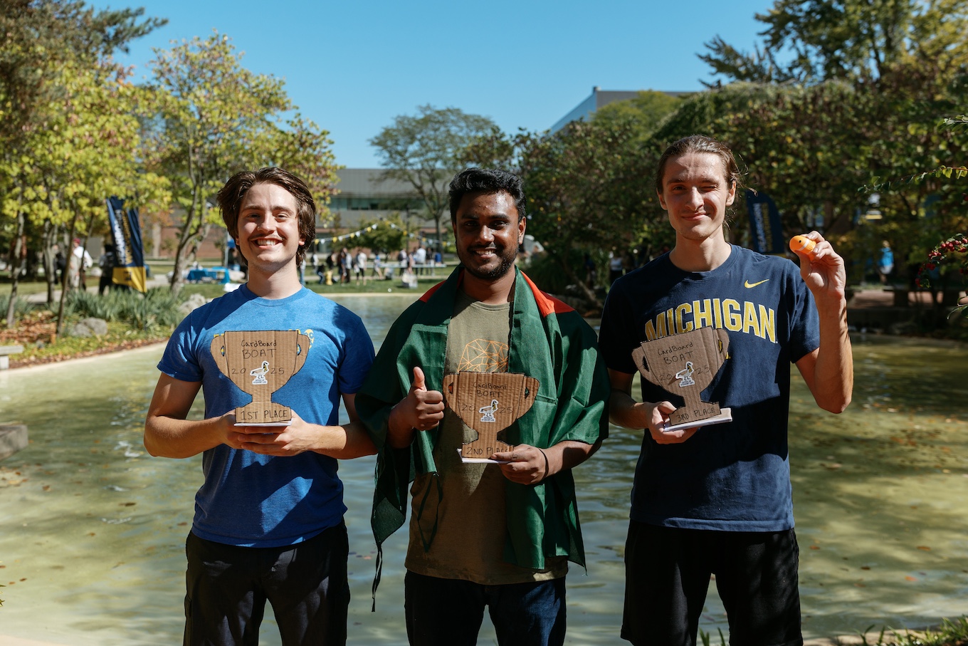 Near a pond, three students hold trophies after winning the cardboard boat races.