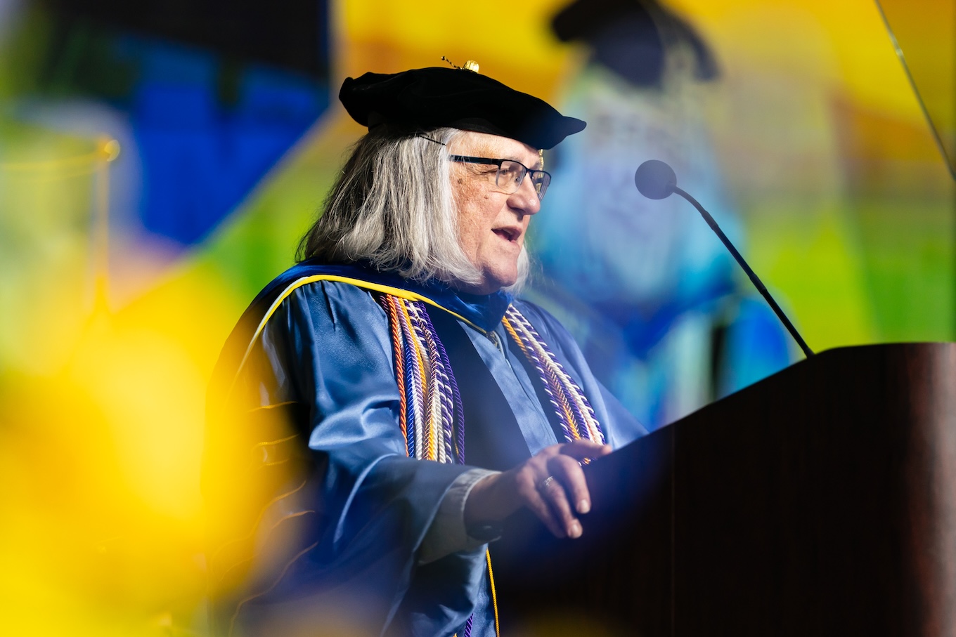 A man with medium-length gray hairs is wearing a black doctoral graduation hat. He is speaking into a microphone at a podium