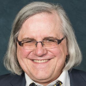 Close-up portrait of an older adult with shoulder-length gray hair, wearing a dark suit jacket, white shirt, and patterned tie.
