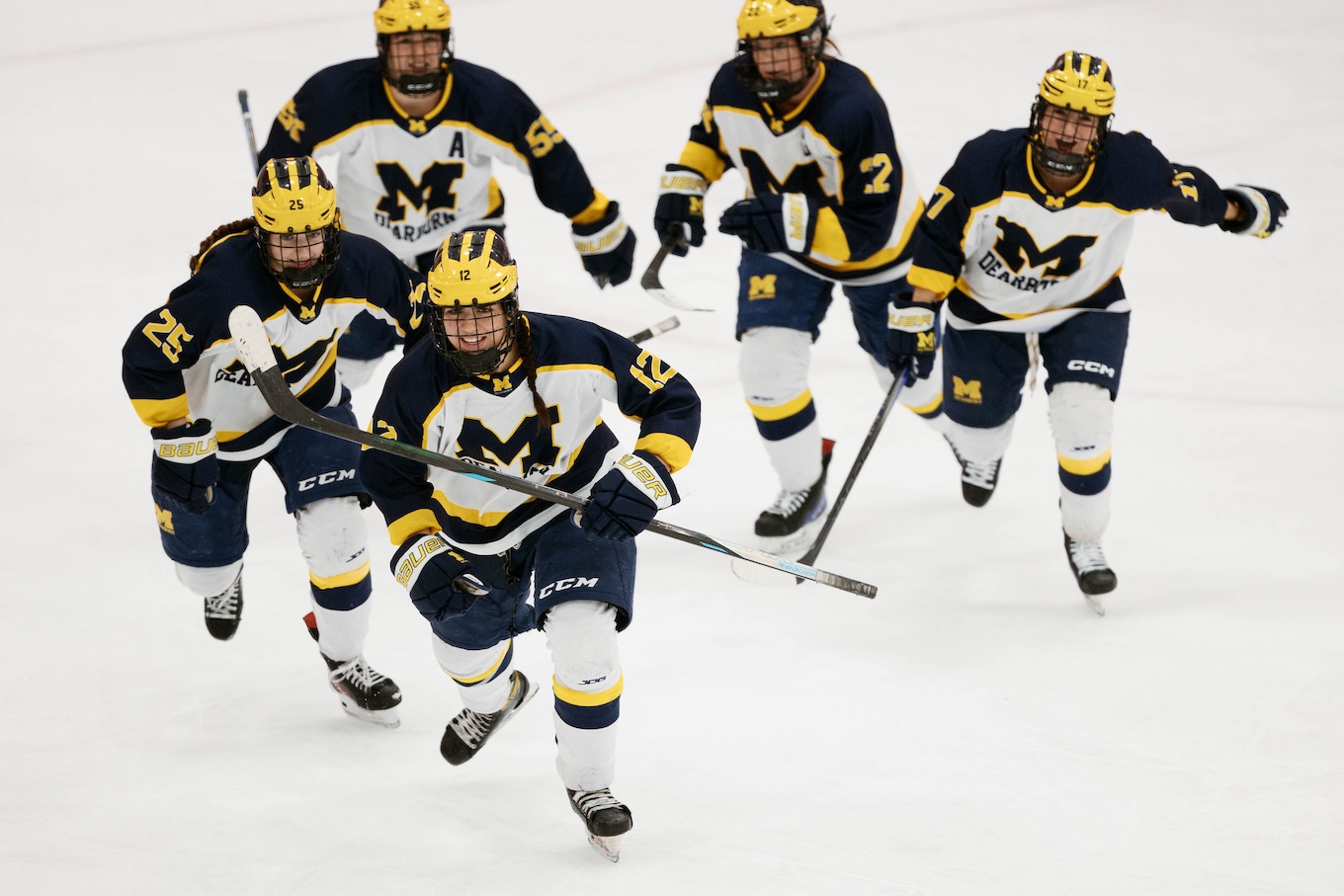 Five female hockey players wearing UM-Dearborn uniforms skate on the ice after scoring a goal.