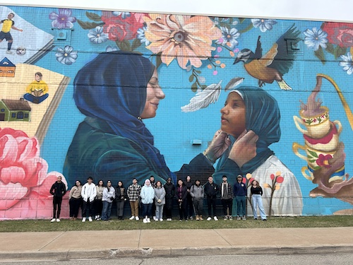 Dr. Carmel Price and students pose in front of a beautiful, hand-painted mural on the side of a building.