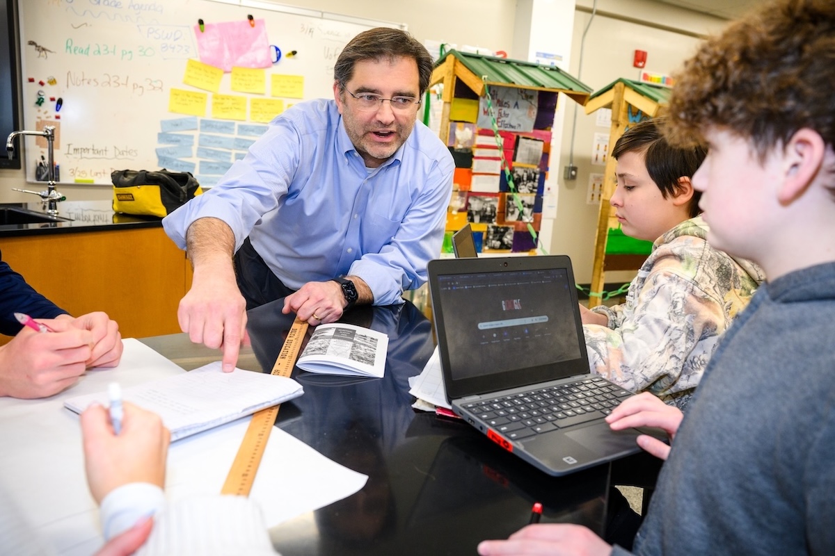 A male teacher talks with two students while they look up information on their laptops