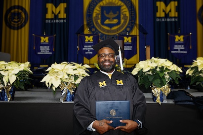 Graduate in cap and gown holding a diploma with University of Michigan-Dearborn banners and poinsettia plants in the background