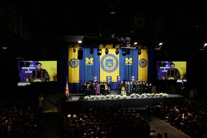 University of Michigan Dearborn commencement ceremony stage with faculty and graduates, blue and yellow banners, and large screens displaying a speaker.