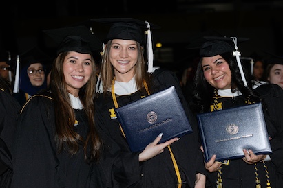 Three graduates in black caps and gowns holding diploma folders with University of Michigan-Dearborn insignia