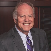 Henry Maier wearing a gray suit, white shirt, and purple striped tie smiles warmly in a professional indoor portrait.