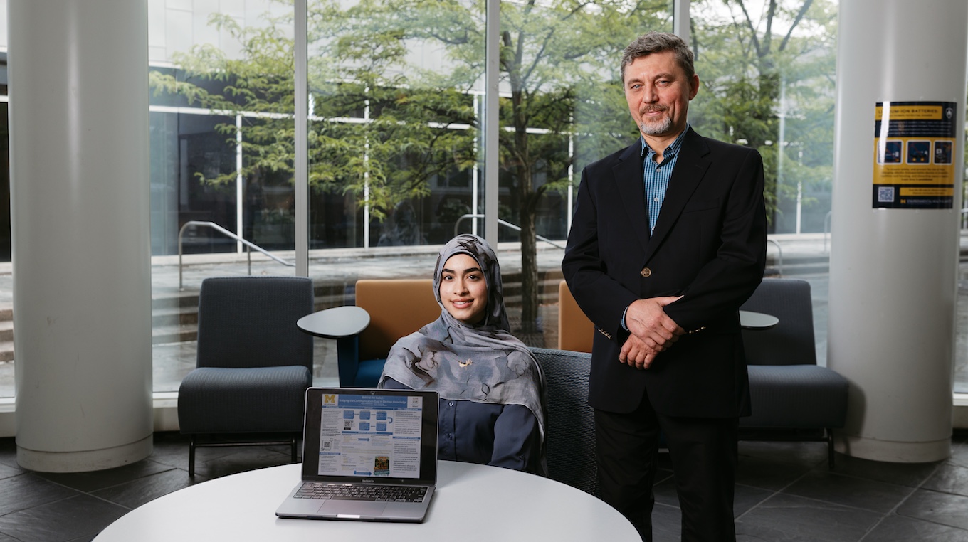 Two people, one seated and one standing, in front of large windows. Amany Alhashidi and Associate Professor of Political Science Vadym Pyrozhenko