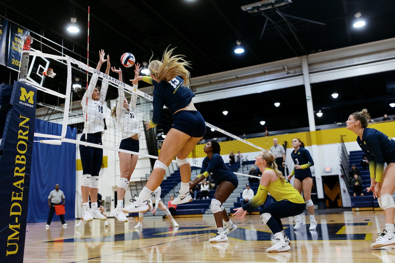 Females play volleyball. One hits a ball over a net while two on the other side of the net work to block it.