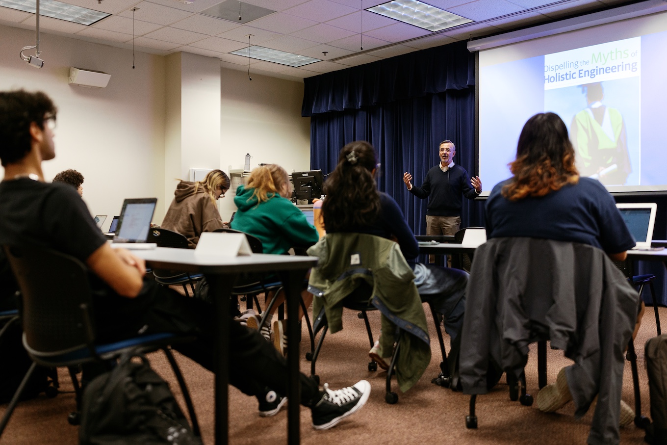 A group of seated students look to the front of the room, where a male professor in a suit stands.