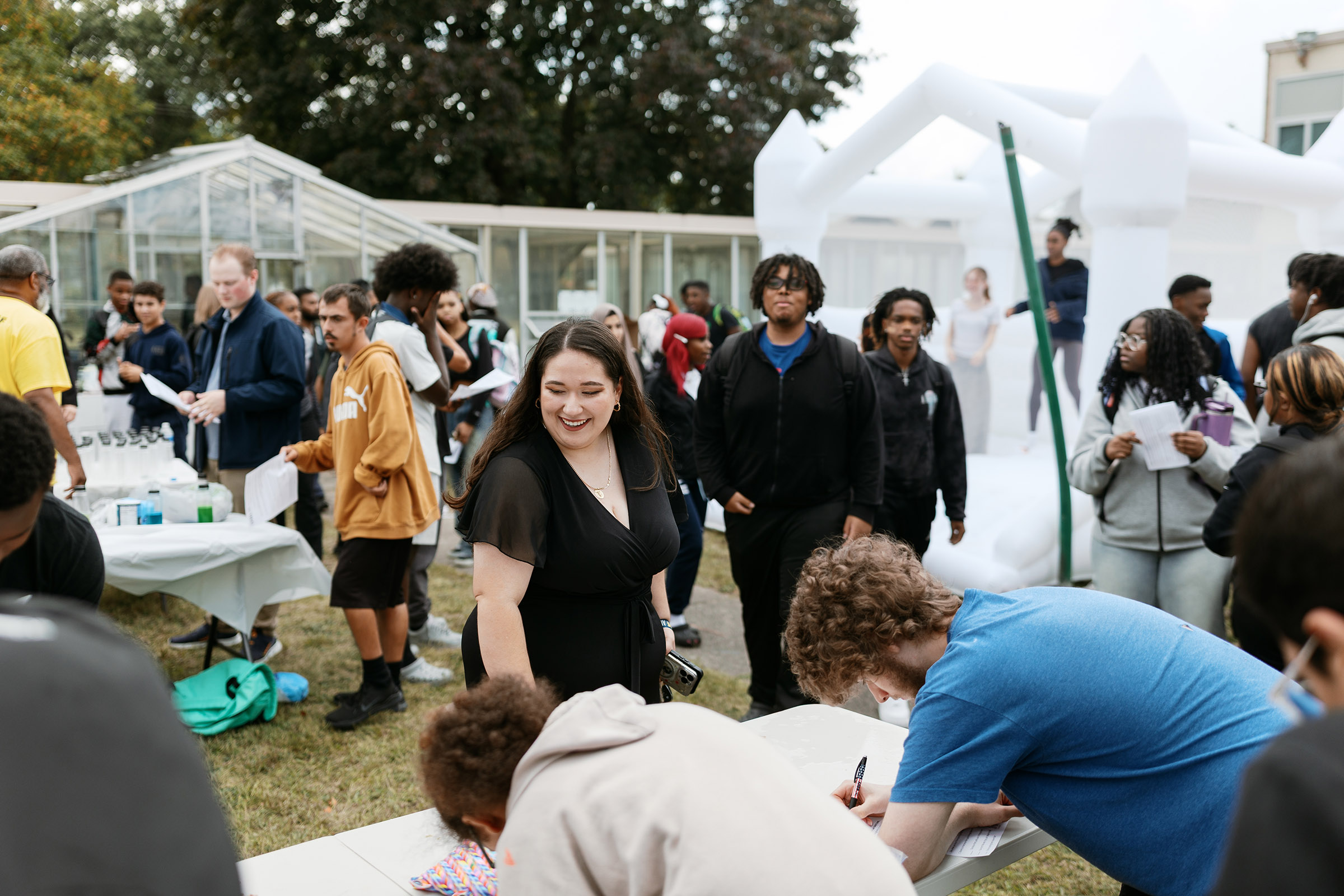 A young woman smiles as she watches students sign up  at an outdoor tabling event at a high school