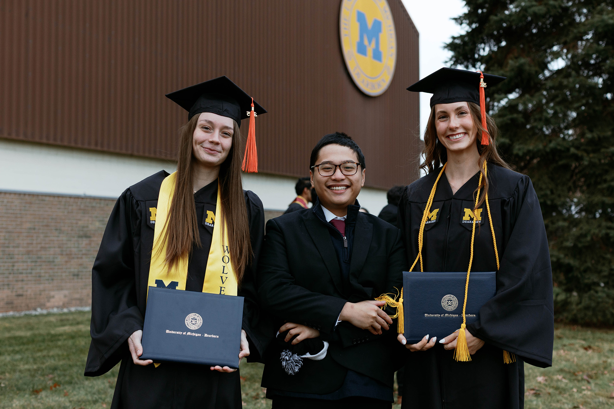 Two female students in graduation robes stand with their male friend while posing for an outdoor portrait on graduation day