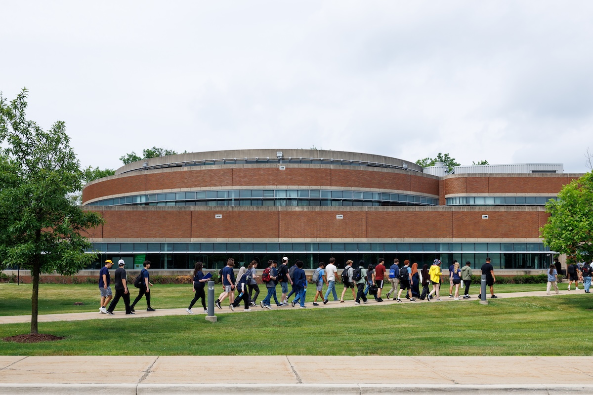 A long line of students walks along a concrete pathway in front of a large, curved brick campus building surrounded by green lawns.