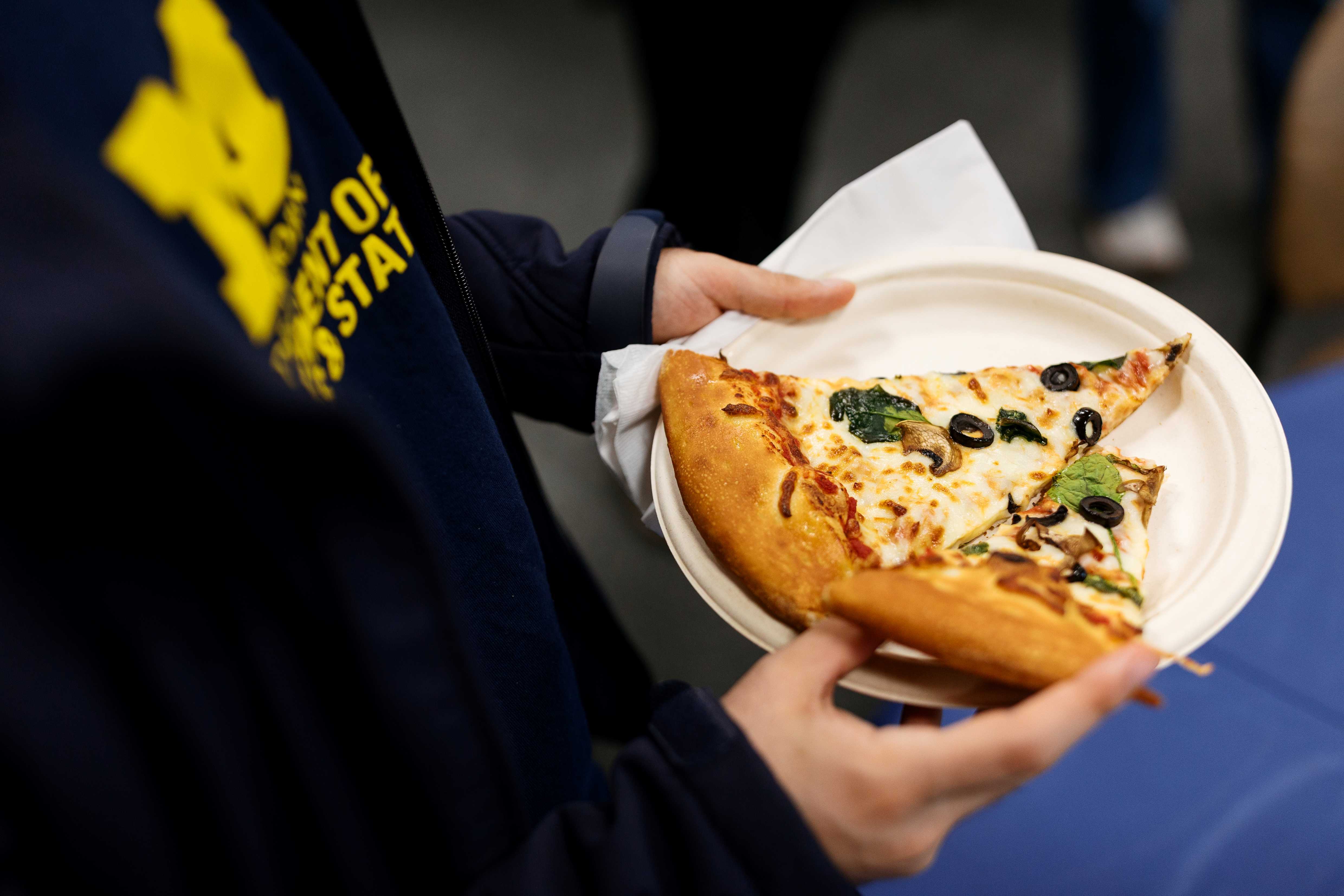 Hands hold a paper plate with two slices of pizza topped with olives and spinach.A napkin is tucked under the plate at an indoor gathering.A navy University of Michigan sweatshirt is visible in the foreground.