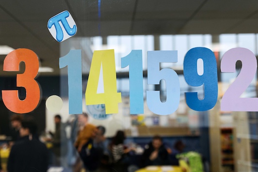 Colorful paper cutouts of “3.141592” are displayed on a glass window, with a pi symbol above.An indoor lounge or campus common area is visible through the glass.Groups of people sit and talk in the blurred background.