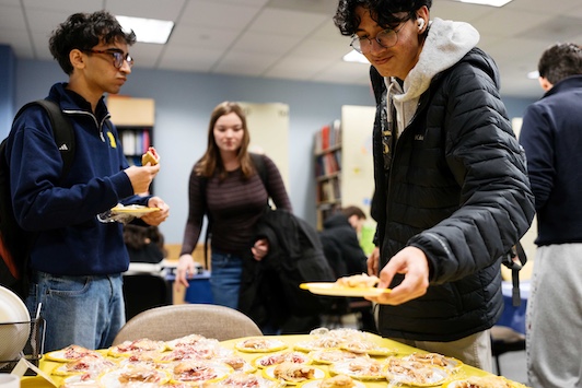 Students stand and chat at an indoor campus event with snacks.A table in the foreground is covered with plates of pastries and cookies wrapped in plastic.People carry plates and backpacks in a room with bookshelves and fluorescent lighting.
