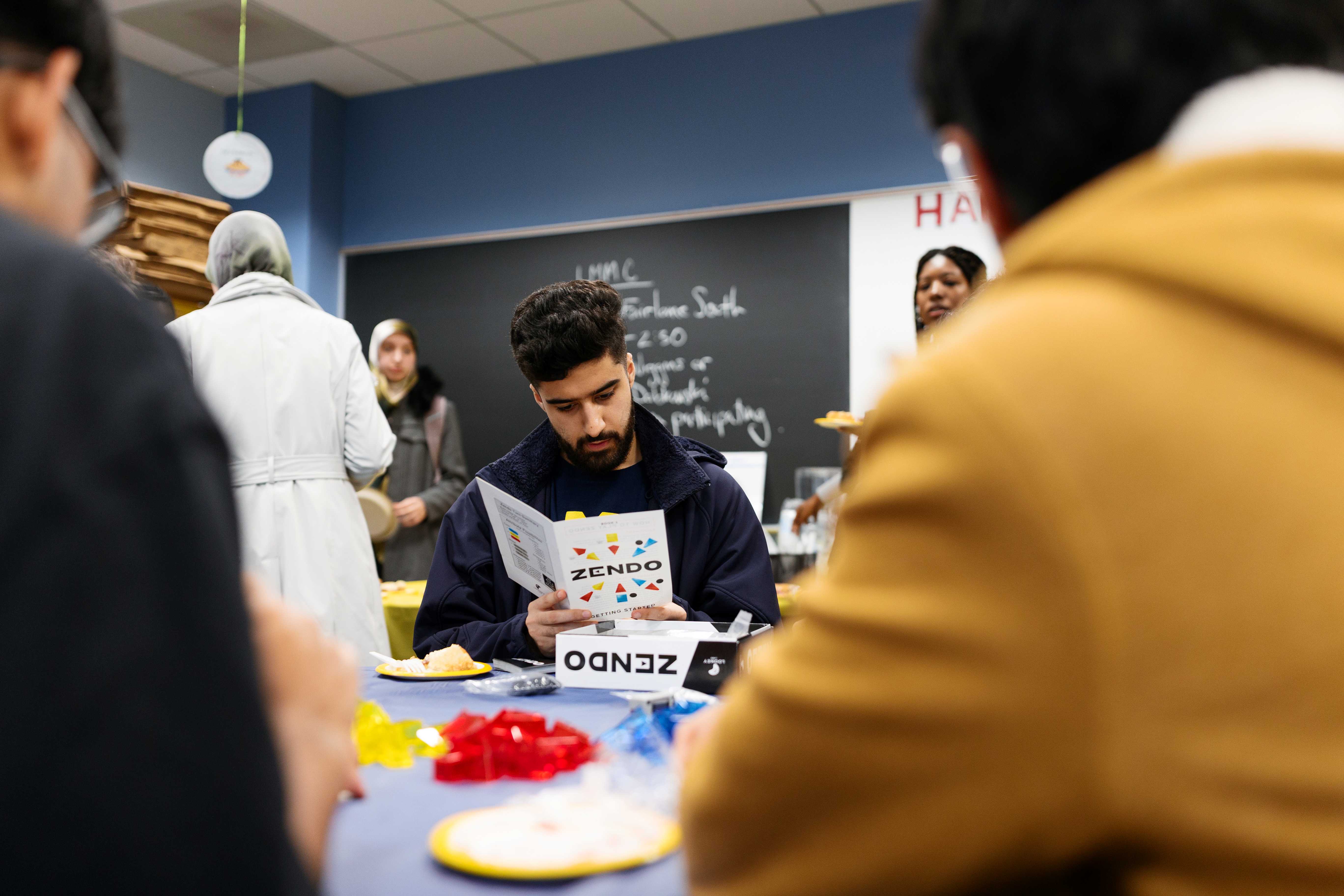 Students gather around a table in a campus lounge during a game night.One person reads a “Zendo” game booklet, with the game box and pieces on the table.Plates of snacks and cups sit nearby, with a chalkboard and other attendees in the background.