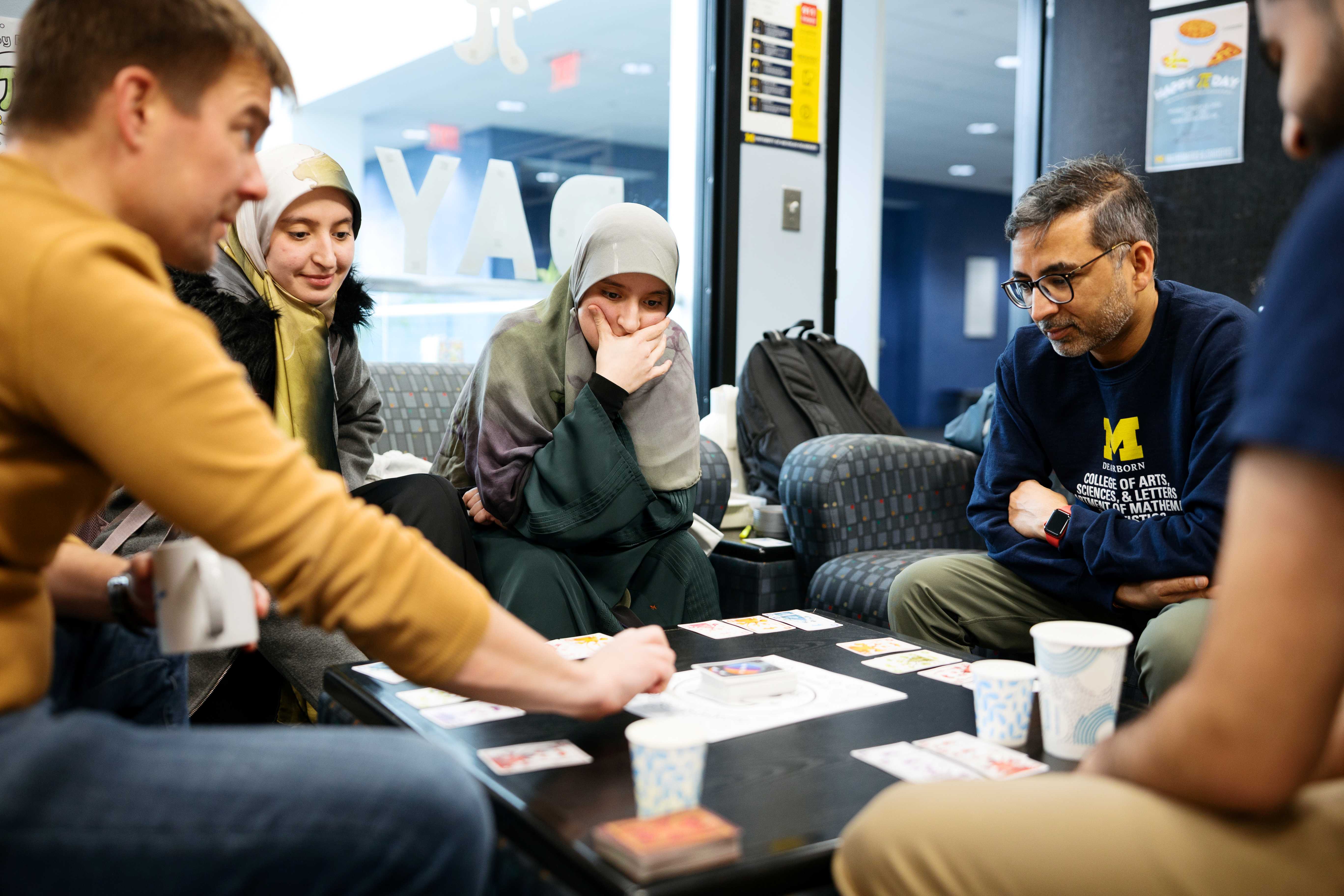 Faculty & students sit in a lounge around a low table, playing a card game.Paper cups, cards, and a small deck box are spread across the tabletop.A person in a University of Michigan sweatshirt leans in, watching the game.