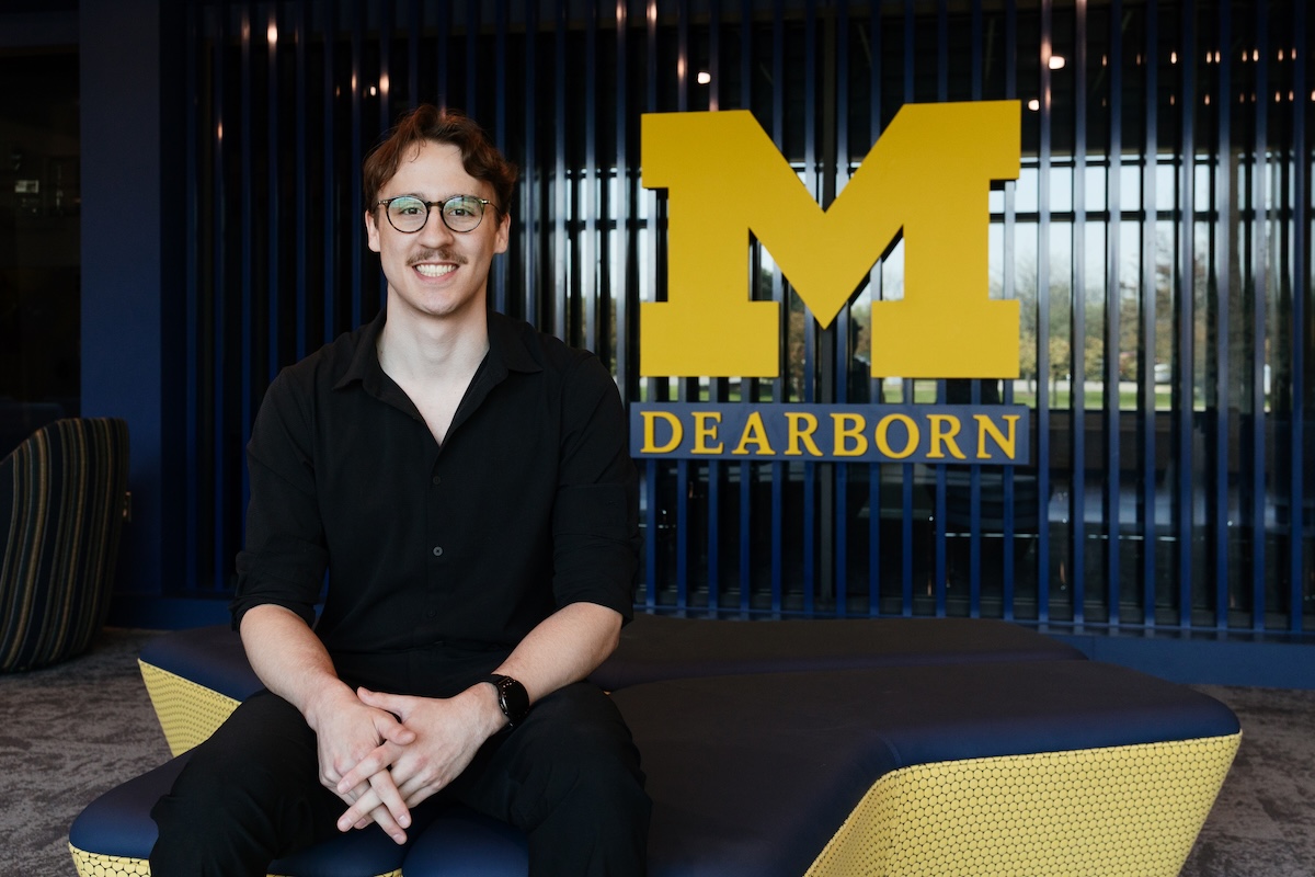 A young man with glasses, a mustache, and a black button-down shirt smiling while sitting on a bench in front of a University of Michigan-Dearborn sign.