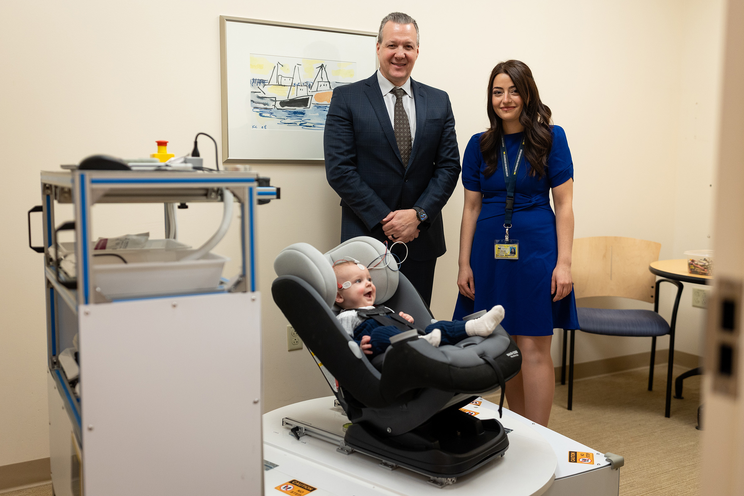 A male doctor and a female doctor pose for a photograph, standing behind a 10-month-old baby sitting in a specialized medical device