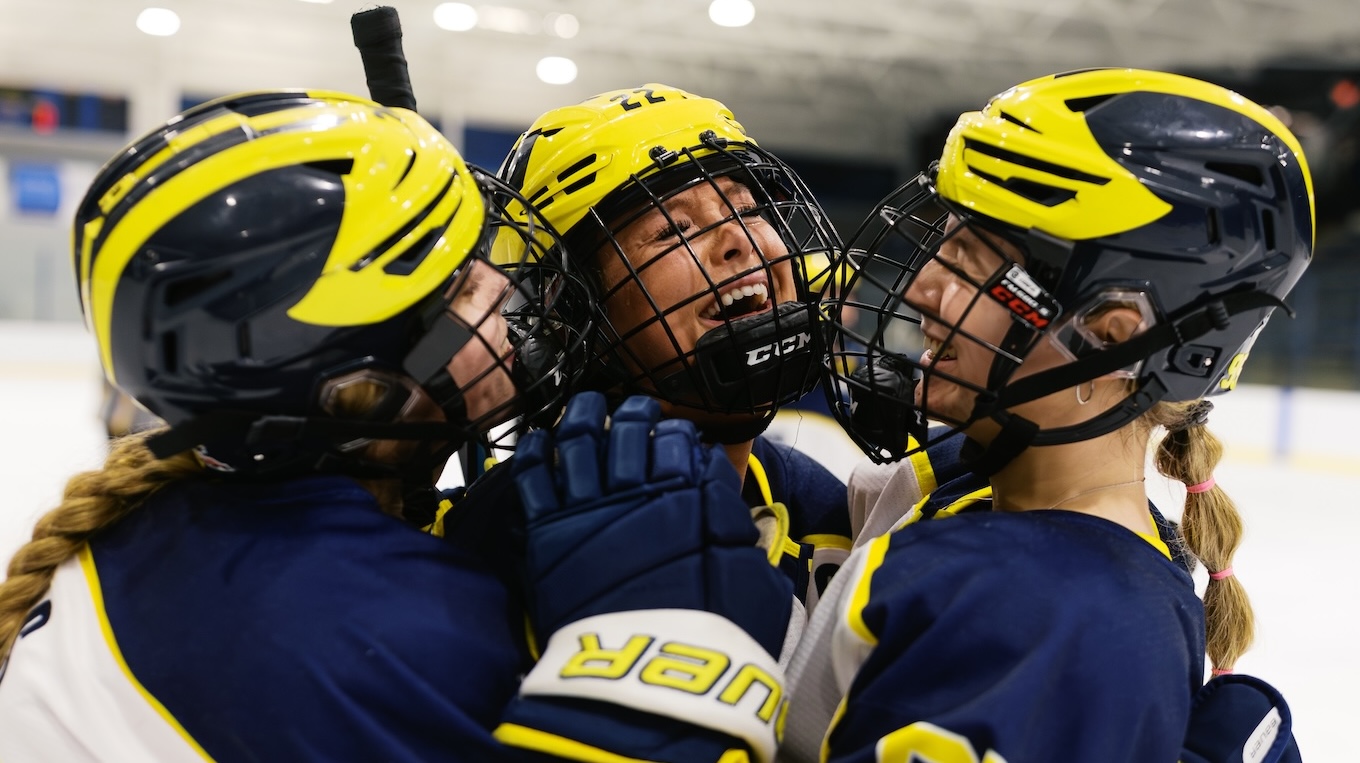 Three female hockey players hug on the ice after a goal is scored.
