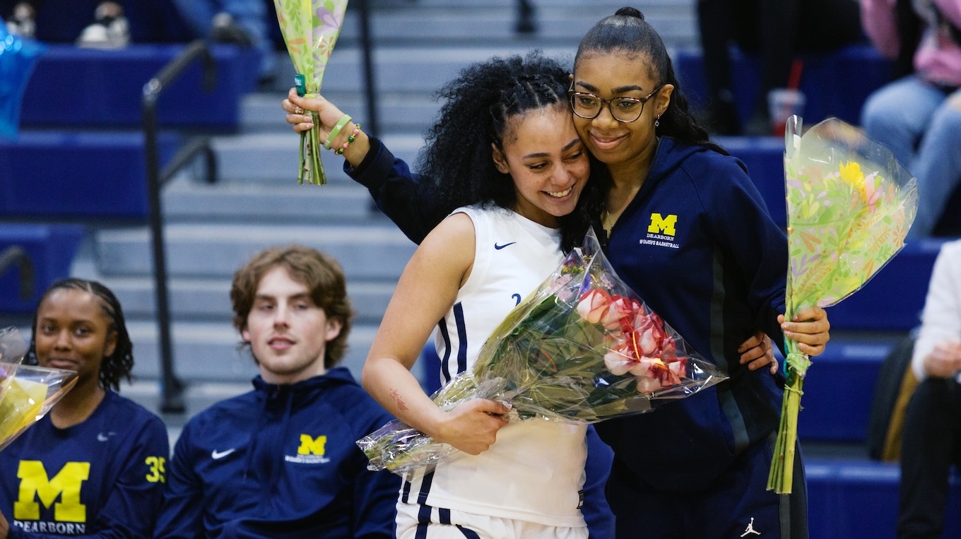Two women holding flowers hug each other on the sidelines at the UM-Dearborn women's basketball game.