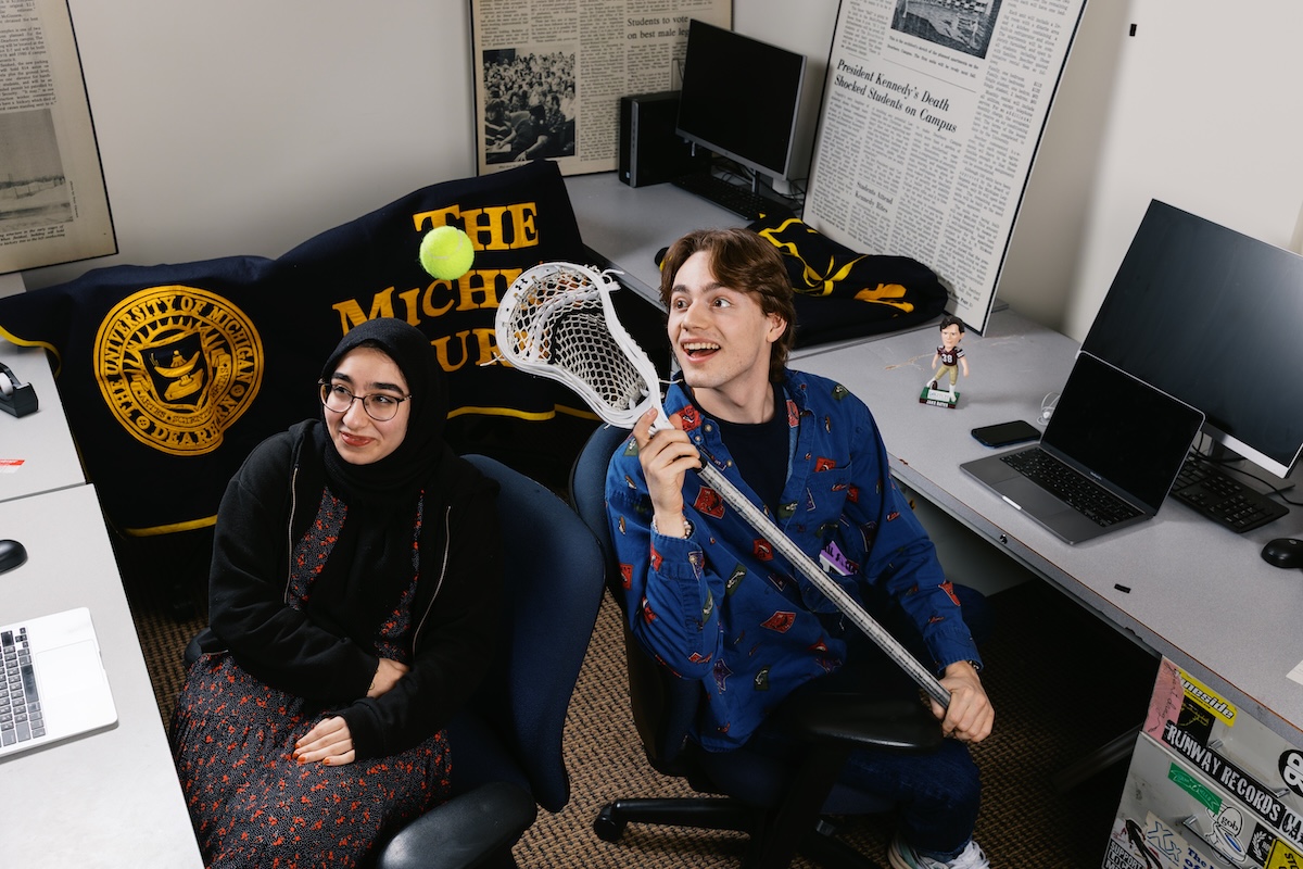 Two people - a female and male - sit in the Michigan Journal office. She smiles and he bounces a tennis ball with a lacrosse stick
