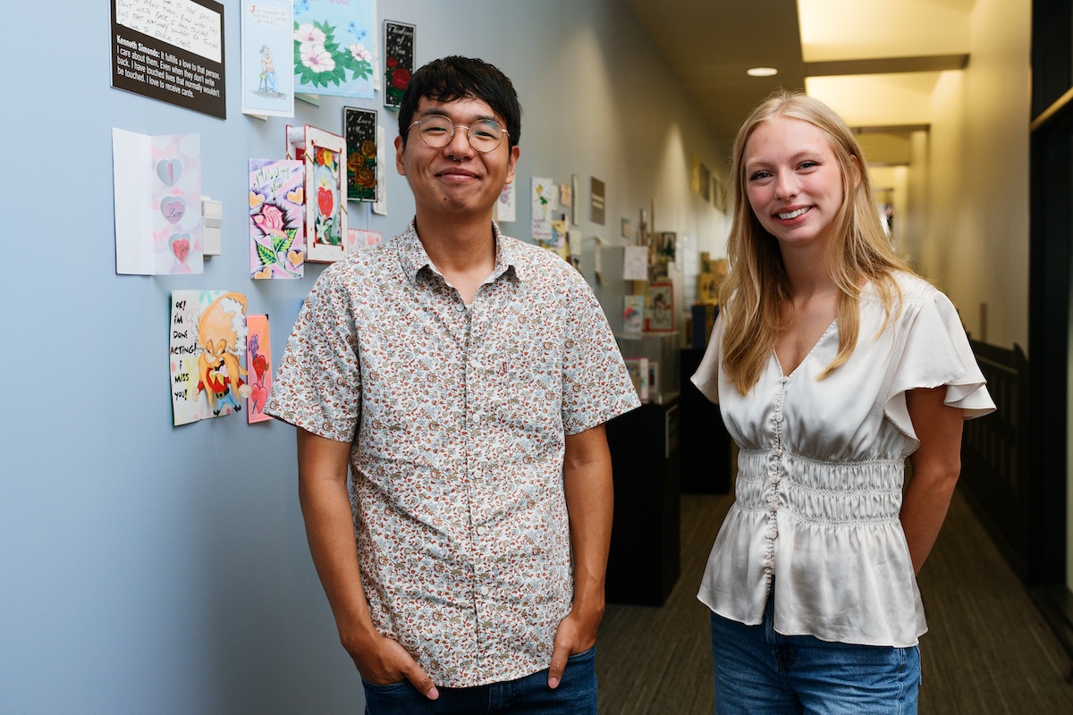 A man with glasses and a patterned short-sleeve button-down shirt stands next to a blonde woman in a white flutter-sleeve top and blue jeans, both smiling at the camera in a hallway with handmade cards displayed on the blue wall behind them.