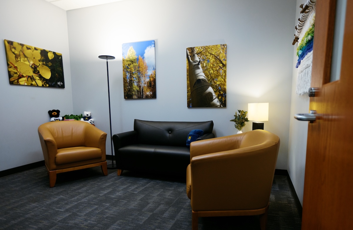 A cozy, quiet interview room furnished with two tan armchairs, a dark sofa, floor and table lamps, and nature photography prints on light gray walls.