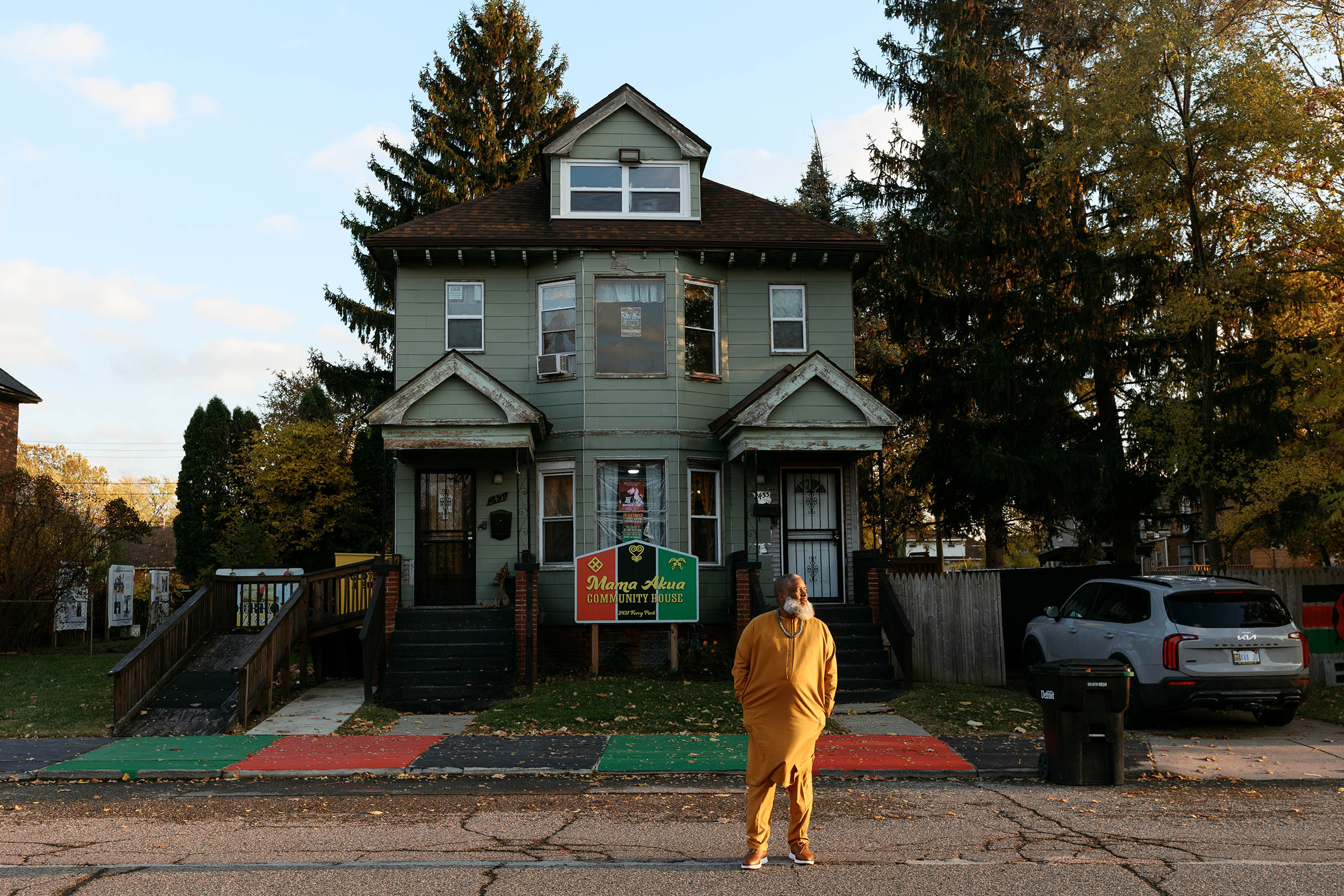 A man in a yellow suit stands outside a green two-story house in a Detroit neighborhood on a late fall day